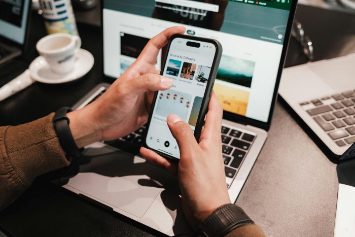 A man's hands holding a cell phone looking at  photos on the phone with  a laptop on the desk in front of him in the background.